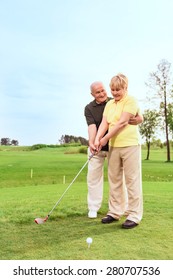 Teaching With Love. Old Man Standing On Course Behind His Wife And Teaching Her To Play Golf 