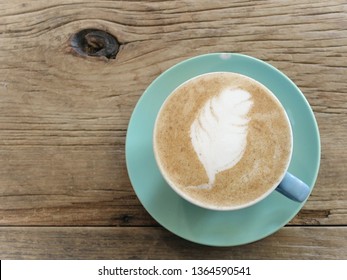 A Teal Colored Cup Of Coffee With Oat Milk And Feather Shaped Latte Art Sits On The Vintage Wooden Table, A Very Hipster Drink For Breakfast Or Long Study Hours.