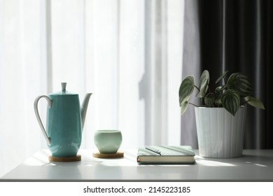 Teal cup and teapot with notebook and on white table with plant pot in front of a see-through curtain - Shutterstock ID 2145223185