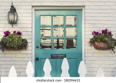A teal wooden front door to a home, with white picket fence gate in foreground. The door is framed by two flower planters, and detail of the white, brick house. 