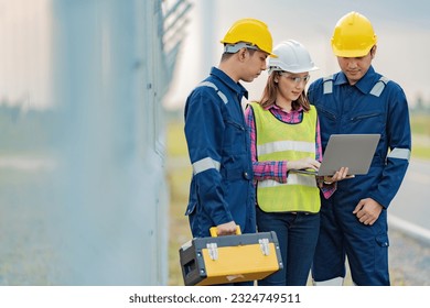 A team of 2 male engineers and an Asian female supervisor working on laptops standing near the fence of an industrial factory. Put on the mechanic's uniform, vest, helmet and toolbox.
