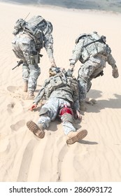 Team of airborne infantry paratroopers saving life of injured brother in arms dragging carrying him on desert sand. No man left behind