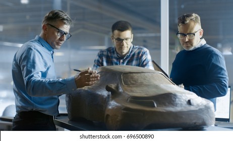 Team Of Automotive Design Engineers Discusses New Prototype Model Made Of Plasticine Clay. They Work In A Large Car Factory.