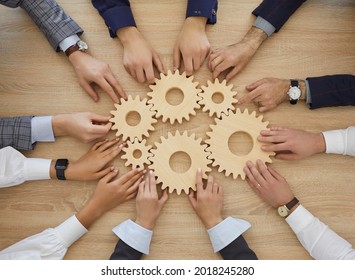 Team Of Business People Join Gearwheels. High Angle, Overhead View Of Circle Of Hands Holding Cogs On Office Table. Metaphor For Good Effective Business System, Cooperation, Teamwork And Efficiency