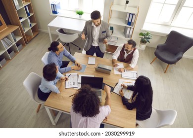 Team Of Businesspeople Having A Discussion In A Meeting. Group Of Young And Mature Business Teammates Sitting Around Table In Office Interior And Listening To Manager. View From Above, High Angle Shot