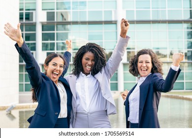 Team of cheerful businesswomen raising hands. Group of happy multiethnic female colleagues triumphing and smiling at camera on street. Success concept