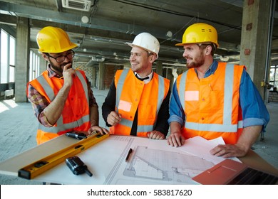 Team of cheerful construction workers discussing project details with executive supervisor standing at table with blueprints, tools and laptop on it