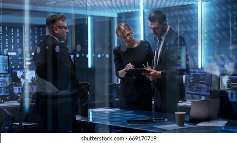 Team Of Chief Engineers Have Emergency Meeting In System Controls Center. They Consult Data On Tablet Computer And Analyse It. Computers In The Room Have Various Information On Their Screens.