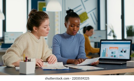 Team Of Colleagues Analzying Market Share Looking At Laptop Comparing Sales Charts On Clipboard With Business Papers On Desk. Startup Employee Working With African American Coworker On Group Project.