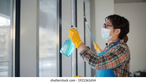 Team Of Commercial Cleaners In Safety Mask Working In Office Cleaning Windows. Side View Of Diverse Janitors Wearing Uniform And Protective Mask Wiping Window With Detergent Sprayer And Cloth