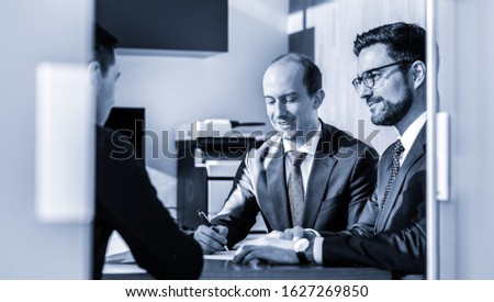 Team of confident successful business people reviewing and signing a contract to seal the deal at business meeting in modern corporate office. Business concept. Greyscale blue toned image.
