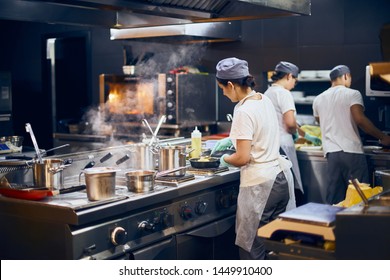 The Team Of Cooks Backs In The Work In The Modern Kitchen, The Workflow Of The Restaurant In The Kitchen. Copy Space For Text