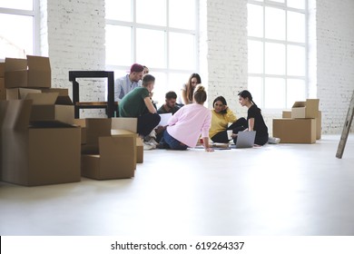 Team Of Coworkers Working Together On Startup Project Just Moved In New Office Sitting Near Carton Boxes With Stuff And Goods, Male And Female Crew Members Planning How To Arrange Space With Furniture