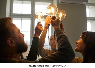 Team of creative minds. Group of smiling intelligent young and senior people holding and raising up bright, shining, glowing Edison light bulbs as symbol of developing collective ideas and innovations - Shutterstock ID 2079249214