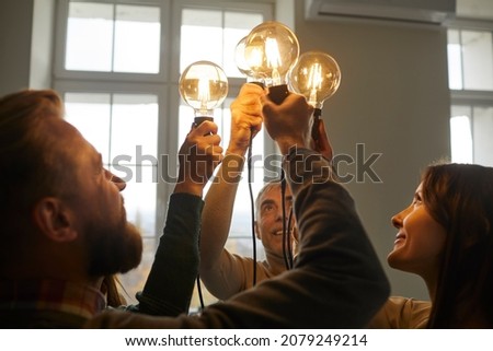 Team of creative minds. Group of smiling intelligent young and senior people holding and raising up bright, shining, glowing Edison light bulbs as symbol of developing collective ideas and innovations