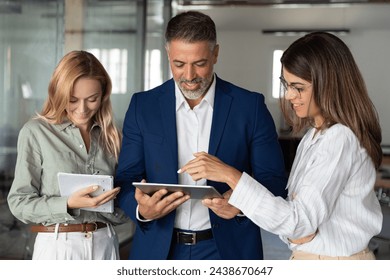 Team of diverse partners mature Latin businessman and European businesswoman discussing project on tablet standing in office. Colleagues, professional businesspeople man and two woman working together - Powered by Shutterstock