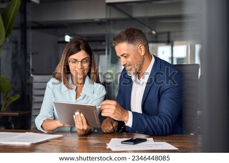 Team of diverse partners mature Latin business man and European business woman discussing project on tablet sitting at table in office. Two colleagues of professional business people working together.