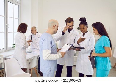 Team Of Doctors And Nurses Standing In Hospital Staff Room And Discussing Difficult Problem. Group Of Coworkers In Scrubs And Lab Coats Studying Report Files Or Complicated, Interesting Medical Case