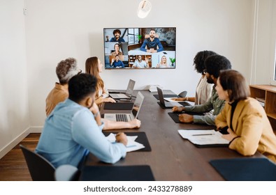 Team engaged in a virtual meeting with a display screen, showing both remote participants and in-person colleagues interacting seamlessly, integration of modern technology in collaborative workspaces - Powered by Shutterstock