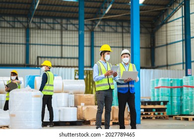 Team Engineers And Foreman Wear A Mask, Hard Hat, And Vest. Standing, Consult Discuss Industrial Production Management Through Data Files With In Mask Factory. Employees Working In The Background.