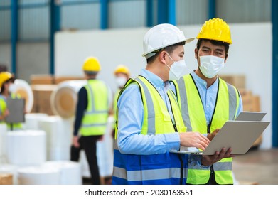 Team Engineers And Foreman Wear A Mask, Hard Hat, And Vest. Standing, Consult Discuss Industrial Production Management Through Data Files With In Mask Factory. Employees Working In The Background.