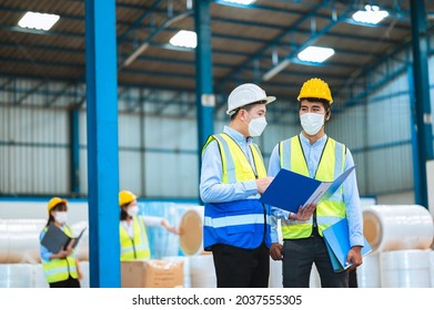 Team Engineers And Foreman Wear A Mask, Hard Hat, And Vest. Standing, Consult Discuss Industrial Production Management Through Data Files With In Mask Factory. Employees Working In The Background.