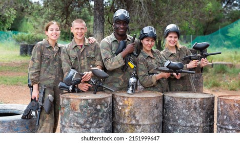 Team of excited friends paintball players in camouflage standing with guns on paintball playing field outdoors
