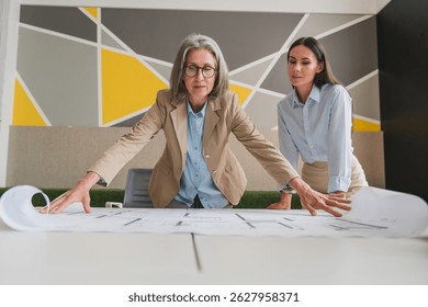 Team of female architects working on construction plans in meeting room. Engineers discussing on project in office. Businesswoman and her colleague standing around table working on blueprint.