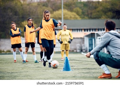 Team of female soccer players practicing with their coach on playing field. Focus is on woman dribbling ball among cones. - Powered by Shutterstock