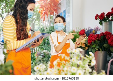 Team Of Florists In Medical Masks Discussing Which Flowers To Order For The Next Week And Filling Document