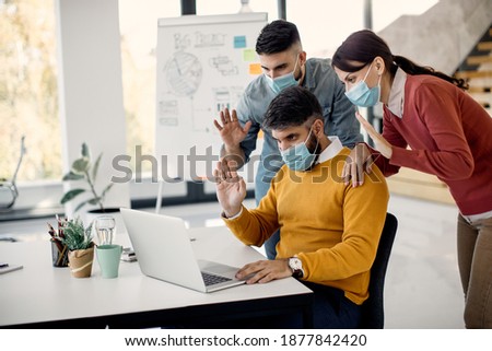 Team of freelance workers wearing protective face masks and waving during video call over laptop in the office. 