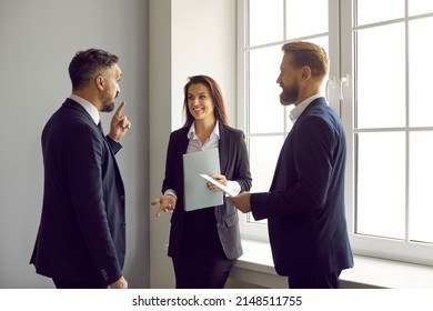 Team Of Happy Business People Having A Discussion. Group Of Three Coworkers Standing Together By The Window And Talking. Smiling Man And Woman Having A Conversation With A More Experienced Colleague