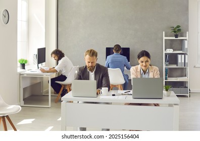 Team Of Happy Business People Working On Computers. Group Of Busy Corporate Employees Sitting At Office Tables And Working On Modern Laptop And Desktop PCs