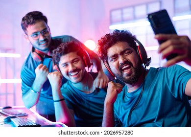 Team Of Happy Cheerful Professional Gamers With Headphones Vlogging, Live Streaming, Or Taking Selfie On Mobile Phone In Front Of Monitors During Esports Video Gaming Tournament