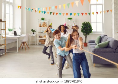 Team of happy smiling diverse children in funny cone hats playing tug-of-war in spacious decorated room at birthday party at home. Group of kids enjoying exciting games in after school club or center