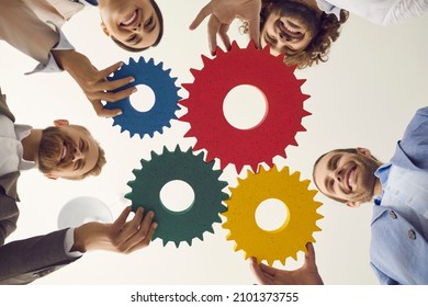 Team of happy young business people joining colorful green, blue, red and yellow cog wheels as symbol or metaphor for teamwork and developing new effective strategy. View from below, low angle shot