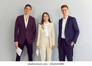Equipo de jóvenes empresarios felices parados en la oficina. Grupo de dos hombres y una mujer con traje elegante, elegante y formal, azul, violeta y beige, posados por una pared gris claro
