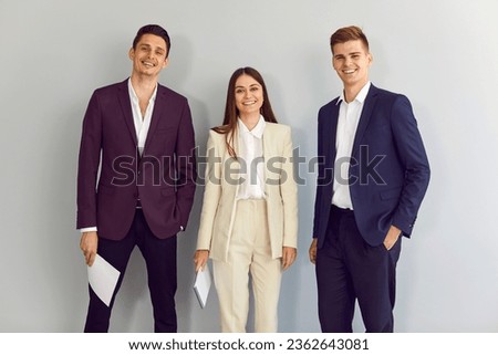Team of happy young business people standing in the office. Group of two men and one woman wearing elegant, smart, formal dress code blue, purple and beige suits posing by a light grey wall