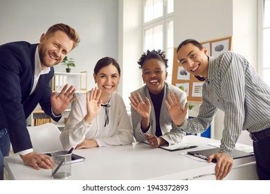 Team of happy young diverse business people sitting and standing around office table, smiling, looking at camera and waving hello, greeting international coworkers in remote work meeting via Internet