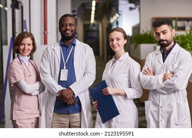 Team Of Happy Young Intercultural Healthcare Workers In Uniform Looking At Camera While Standing In Row In Corridor Of Modern Clinics