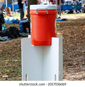 A Team Has Set Up Their Orange Water Cooler Jug On Top Of White Wood Boards In A Park During A Cross Country Runners Race.