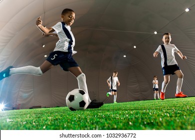 Team Of Intercultural Boys Running Throughout Football Field, One Of Them Kicking The Ball