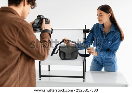 Team of male photographer and smm manager taking photos of stylish handbag on white platform background in modern photostudio, woman helping to make content photoshoot