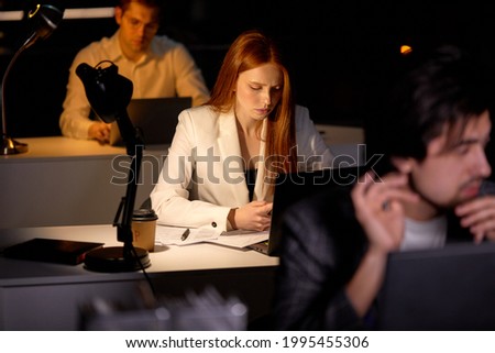 Team of managers office workers working on project very late, using computer. Hardworking people in formal wear engaged in work, thinking, brainstorming, focus on redhead female with smartphone