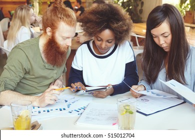 Team Of Marketing Experts Developing Business Strategy At Cafe African Woman Presenting Business Plan To Her Partner With Red Beard On Digital Tablet While Their Asian Colleague Analysing Graphs