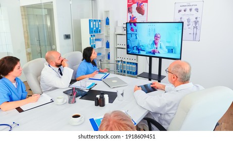 Team Of Medical Staff During Video Conference With Doctor In Hospital Meeting Room. Medicine Staff Using Internet During Online Meeting With Expert Doctor For Expertise, Nurse Taking Notes.