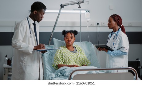 Team Of Medical Workers Doing Consultation With Patient In Hospital Ward Bed. Physician Talking To Sick Person About Healthcare Treatment While Woman Holding Digital Tablet To Help