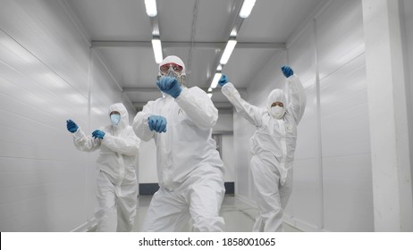 Team Of Medical Workers In Hazmat Suits Dancing And Walking In Hospital Corridor. Funny Disinfectors In Respiratory Mask And Protective Uniform Dancing Celebrating End Of Epidemic