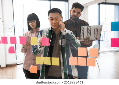 Team members brainstorming creative ideas on transparent board using sticky notes and electronic devices. Professional collaboration and planning in modern office environment - Powered by Shutterstock