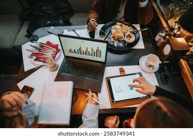 Team members collaborating, analyzing data and charts from laptops and tablets, discussing ideas over coffee and snacks in a cafe setting, fostering teamwork and creativity.